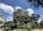 A modern, wavy building rises behind lush green trees, set against a bright blue sky dotted with white clouds. The architecture is surrounded by urban elements and a well-maintained park area, creating a contrast between nature and urban design.