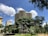 A modern, wavy building rises behind lush green trees, set against a bright blue sky dotted with white clouds. The architecture is surrounded by urban elements and a well-maintained park area, creating a contrast between nature and urban design.