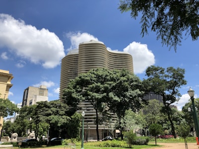 A modern, wavy building rises behind lush green trees, set against a bright blue sky dotted with white clouds. The architecture is surrounded by urban elements and a well-maintained park area, creating a contrast between nature and urban design.
