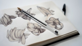 A sketchbook filled with detailed black and white pencil sketches of abstract floral designs. A pen is resting diagonally across the open pages, suggesting a moment paused in creativity. The background is minimalist, emphasizing the artwork.
