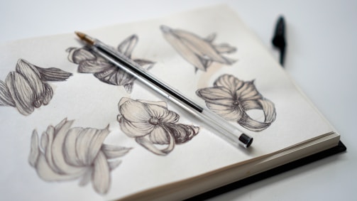A sketchbook filled with detailed black and white pencil sketches of abstract floral designs. A pen is resting diagonally across the open pages, suggesting a moment paused in creativity. The background is minimalist, emphasizing the artwork.