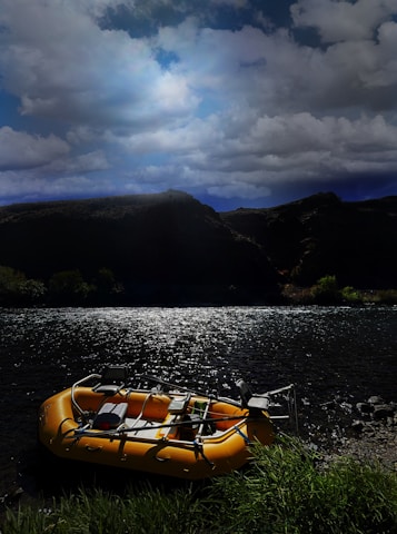 A bright yellow inflatable raft is positioned on the grassy bank of a river, with sunlight reflecting off the water, creating a shimmering effect. The background features a range of hills silhouetted against a dramatic sky filled with large, billowing clouds.