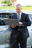 A man in a business suit is standing next to a car while using a laptop. The background includes a grassy area and a building with a garage door. The man appears focused on the task at hand.