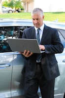 A man in a business suit is standing next to a car while using a laptop. The background includes a grassy area and a building with a garage door. The man appears focused on the task at hand.