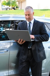 A man in a business suit is standing next to a car while using a laptop. The background includes a grassy area and a building with a garage door. The man appears focused on the task at hand.