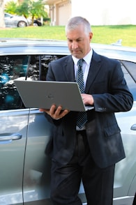 A man in a business suit is standing next to a car while using a laptop. The background includes a grassy area and a building with a garage door. The man appears focused on the task at hand.