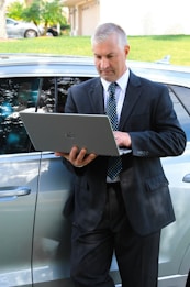 A man in a business suit is standing next to a car while using a laptop. The background includes a grassy area and a building with a garage door. The man appears focused on the task at hand.