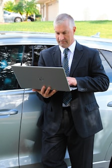 A man in a business suit is standing next to a car while using a laptop. The background includes a grassy area and a building with a garage door. The man appears focused on the task at hand.