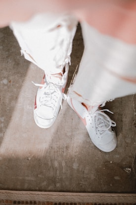 White sneakers with pink accents are worn with ripped, faded jeans. The floor underneath seems to be made of worn concrete with some small imperfections and a hint of sunlight casting a soft shadow.