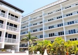 A modern multi-story building with a white facade and numerous balconies, surrounded by lush green palm trees. The structure features clean lines and a symmetric design, indicative of contemporary architecture.