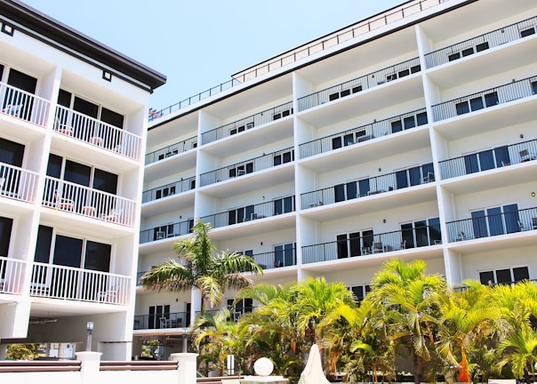 A modern multi-story building with a white facade and numerous balconies, surrounded by lush green palm trees. The structure features clean lines and a symmetric design, indicative of contemporary architecture.