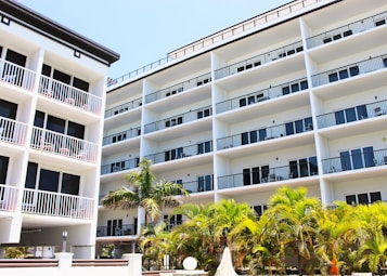 A modern multi-story building with a white facade and numerous balconies, surrounded by lush green palm trees. The structure features clean lines and a symmetric design, indicative of contemporary architecture.