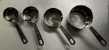 A set of four metallic measuring cups arranged in a line on a textured surface. Each cup is slightly different in size, with handles featuring engraved measurement indicators. The lighting creates reflections on the smooth, shiny surface of the metal.