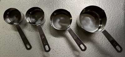 A set of four metallic measuring cups arranged in a line on a textured surface. Each cup is slightly different in size, with handles featuring engraved measurement indicators. The lighting creates reflections on the smooth, shiny surface of the metal.