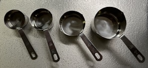 A set of four metallic measuring cups arranged in a line on a textured surface. Each cup is slightly different in size, with handles featuring engraved measurement indicators. The lighting creates reflections on the smooth, shiny surface of the metal.
