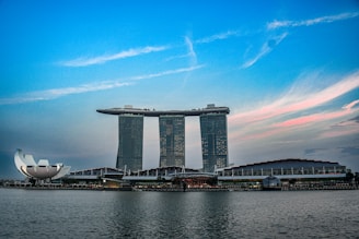 A waterfront cityscape featuring a large, futuristic building with three tower structures connected by a horizontal rooftop section. On the left, a uniquely shaped white structure resembling a lotus flower stands prominently. The sky is a soft gradient of blue with hints of pink clouds as dusk approaches.