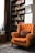 A cozy interior setting with an orange upholstered armchair featuring tufted details and a decorative pillow. Behind the chair is a tall bookshelf filled with various colorful books. A framed picture is hung on the adjacent white wall. To the side, a potted plant sits on a wooden stand, next to a dark, patterned curtain. The floor is covered with an ornate rug, completing the warm and inviting atmosphere.