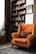 A cozy interior setting with an orange upholstered armchair featuring tufted details and a decorative pillow. Behind the chair is a tall bookshelf filled with various colorful books. A framed picture is hung on the adjacent white wall. To the side, a potted plant sits on a wooden stand, next to a dark, patterned curtain. The floor is covered with an ornate rug, completing the warm and inviting atmosphere.