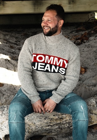 A person is sitting on a log, wearing a gray sweater with a bold logo reading 'Tommy Jeans' and a pair of blue jeans. The background consists of a sandy surface and some wooden beams, possibly part of a rustic structure.
