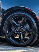 A close-up of a black alloy wheel with a glossy finish. The tire is a high-performance model with visible tread patterns. The surrounding area shows some reflections and a hint of a red brake caliper peeking through the spokes. Part of the car's body is visible, indicating a sleek and modern design.