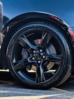 A close-up of a black alloy wheel with a glossy finish. The tire is a high-performance model with visible tread patterns. The surrounding area shows some reflections and a hint of a red brake caliper peeking through the spokes. Part of the car's body is visible, indicating a sleek and modern design.