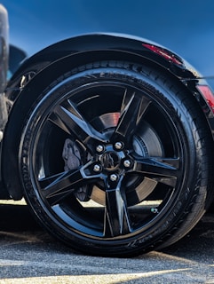 A close-up of a black alloy wheel with a glossy finish. The tire is a high-performance model with visible tread patterns. The surrounding area shows some reflections and a hint of a red brake caliper peeking through the spokes. Part of the car's body is visible, indicating a sleek and modern design.