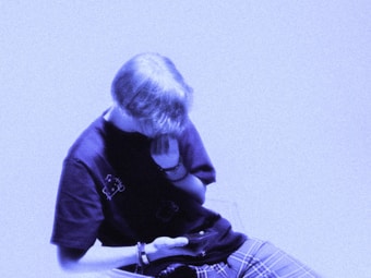 A person with short hair, wearing a dark t-shirt and plaid pants, is sitting and looking down at a smartphone. The image has a bluish tint, creating a calm and subdued atmosphere.