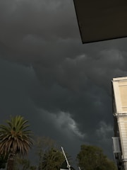Dark storm clouds fill the sky, creating a dramatic and ominous atmosphere. A palm tree and other trees are visible against the backdrop of the looming clouds. The corner of a building is also apparent, with a flagpole nearby.