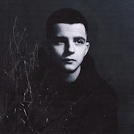 A black and white portrait of a young person, appearing contemplative, with a short haircut and wearing a dark hoodie. Abstract scribbles and marks are present on the left side, adding an artistic and textured element.