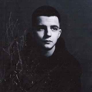 A black and white portrait of a young person, appearing contemplative, with a short haircut and wearing a dark hoodie. Abstract scribbles and marks are present on the left side, adding an artistic and textured element.