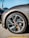 A close-up view of a sleek car wheel with a stylish alloy rim. The tire is branded and mounted on the vehicle, which appears to be parked on asphalt. The car's body is visible with a glossy gray finish, and the background shows part of another vehicle.