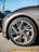 A close-up view of a sleek car wheel with a stylish alloy rim. The tire is branded and mounted on the vehicle, which appears to be parked on asphalt. The car's body is visible with a glossy gray finish, and the background shows part of another vehicle.