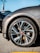 A close-up view of a sleek car wheel with a stylish alloy rim. The tire is branded and mounted on the vehicle, which appears to be parked on asphalt. The car's body is visible with a glossy gray finish, and the background shows part of another vehicle.