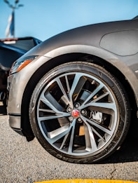 A close-up view of a sleek car wheel with a stylish alloy rim. The tire is branded and mounted on the vehicle, which appears to be parked on asphalt. The car's body is visible with a glossy gray finish, and the background shows part of another vehicle.