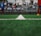 A close-up view of a green soccer field's artificial turf, focusing on a white dividing line running down the center. In the blurred background, there are players wearing sports uniforms and spectators seated in stands.
