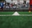 A close-up view of a green soccer field's artificial turf, focusing on a white dividing line running down the center. In the blurred background, there are players wearing sports uniforms and spectators seated in stands.