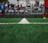 A close-up view of a green soccer field's artificial turf, focusing on a white dividing line running down the center. In the blurred background, there are players wearing sports uniforms and spectators seated in stands.