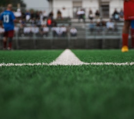 A close-up view of a green soccer field's artificial turf, focusing on a white dividing line running down the center. In the blurred background, there are players wearing sports uniforms and spectators seated in stands.