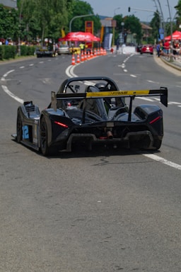 A black racing car with the number 32 on its side is driving down a street that is temporarily converted into a race track. The road is lined with orange traffic cones, and there are spectators on both sides with red tents and greenery visible in the background. The car is sleek and has a large rear wing. The weather appears to be clear and sunny.