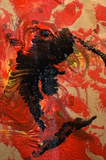 The image consists of an abstract painting with thick, textured strokes of vibrant colors. Predominantly, shades of red and black dominate the canvas, with hints of yellow and orange in the mixture. The brushstrokes are bold and sweeping, creating a sense of dynamic movement across the surface.
