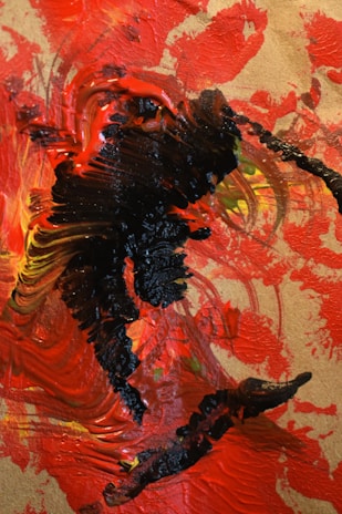 The image consists of an abstract painting with thick, textured strokes of vibrant colors. Predominantly, shades of red and black dominate the canvas, with hints of yellow and orange in the mixture. The brushstrokes are bold and sweeping, creating a sense of dynamic movement across the surface.