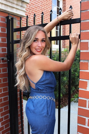 A woman with long, wavy blonde hair wearing a blue sleeveless jumpsuit stands next to a black metal fence. She is smiling and holding onto the top of the fence with her left hand while her right hand grips one of the bars. The background consists of red brick walls and a window.