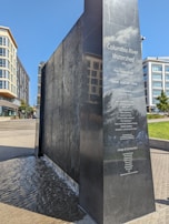 A black granite monument with the title 'Columbia River Watershed' engraved on its surface along with the names of contributors. Water flows gently down one side of the monument. Surrounding the monument are modern buildings with glass windows and greenery in a park-like setting.