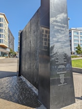 A black granite monument with the title 'Columbia River Watershed' engraved on its surface along with the names of contributors. Water flows gently down one side of the monument. Surrounding the monument are modern buildings with glass windows and greenery in a park-like setting.