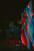 A DJ wearing a red cap is performing at an event, standing behind a laptop and sound equipment. The scene is lit with vibrant neon lights, and an abstract digital display with geometric patterns is visible next to the DJ. The environment appears dark, highlighting the colorful electronic visuals.