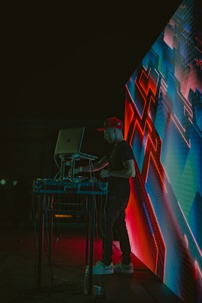 A DJ wearing a red cap is performing at an event, standing behind a laptop and sound equipment. The scene is lit with vibrant neon lights, and an abstract digital display with geometric patterns is visible next to the DJ. The environment appears dark, highlighting the colorful electronic visuals.