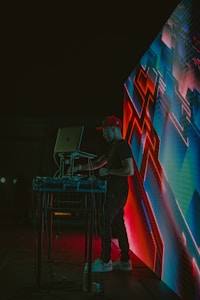 A DJ wearing a red cap is performing at an event, standing behind a laptop and sound equipment. The scene is lit with vibrant neon lights, and an abstract digital display with geometric patterns is visible next to the DJ. The environment appears dark, highlighting the colorful electronic visuals.