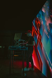 A DJ wearing a red cap is performing at an event, standing behind a laptop and sound equipment. The scene is lit with vibrant neon lights, and an abstract digital display with geometric patterns is visible next to the DJ. The environment appears dark, highlighting the colorful electronic visuals.