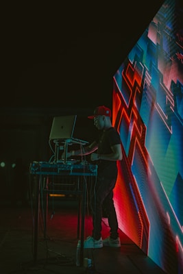 A DJ wearing a red cap is performing at an event, standing behind a laptop and sound equipment. The scene is lit with vibrant neon lights, and an abstract digital display with geometric patterns is visible next to the DJ. The environment appears dark, highlighting the colorful electronic visuals.