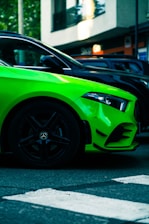 A vibrant green car with sleek, modern lines is parked on a street. The focus is on the front half of the vehicle, highlighting its angular design and glossy finish. The background features an urban setting with a building and additional parked cars.